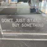 A clear lucite sign is pictured with the words Don't Just Stand There Buy Something engraved in it.