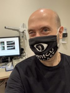 I am pictured sitting on an exam chair with computer monitors behind me. One of them shows the OCT scan. I am wearing an iCantCU Podcast facemask.